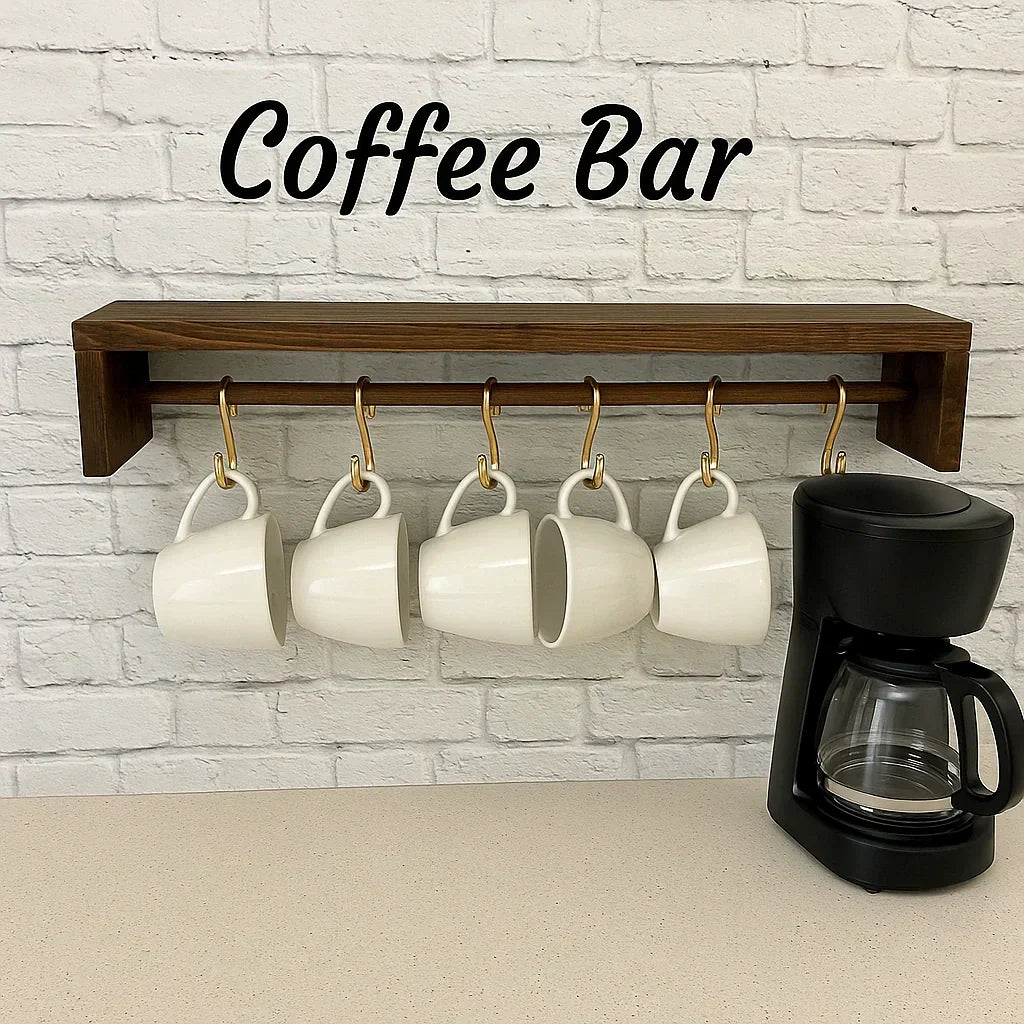 Coffee bar setup with white mugs hanging on a brass rail and wooden shelf, next to a black drip coffee maker on a neutral countertop.