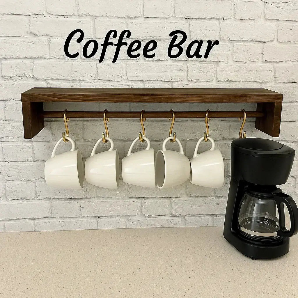 Coffee bar setup with white mugs hanging on a brass rail and wooden shelf, next to a black drip coffee maker on a neutral countertop.