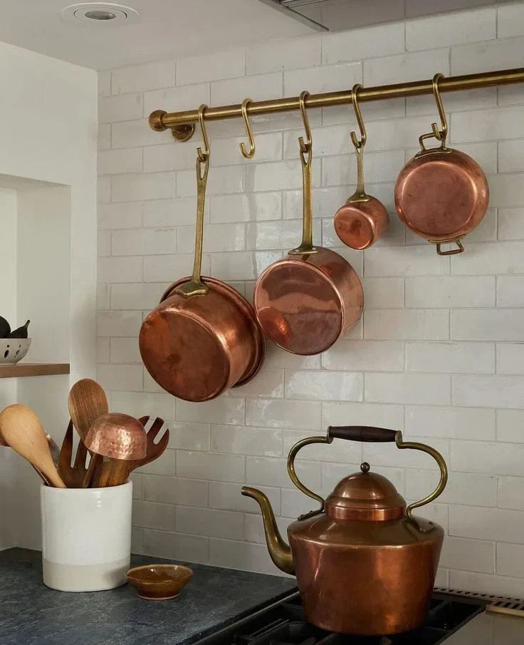 Unlacquered brass pot rack with copper pans and brass S-hooks mounted on white tile kitchen backsplash