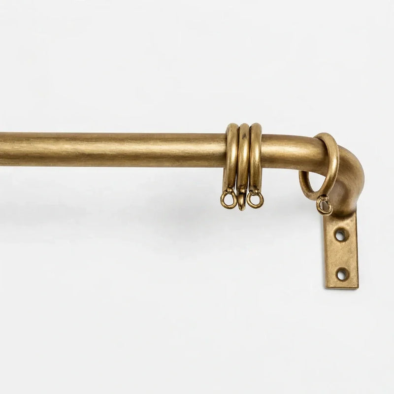 Close-up of an unlacquered brass curtain rod with matching rings and wall-mounted bracket on a matte white background