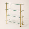 A 4-tier glass shelf bookcase with a polished brass frame, featuring clear glass shelves and round brass finials at the joints and feet, set against a plain off-white background.