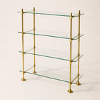 A 4-tier glass shelf bookcase with a polished brass frame, featuring clear glass shelves and round brass finials at the joints and feet, set against a plain off-white background.