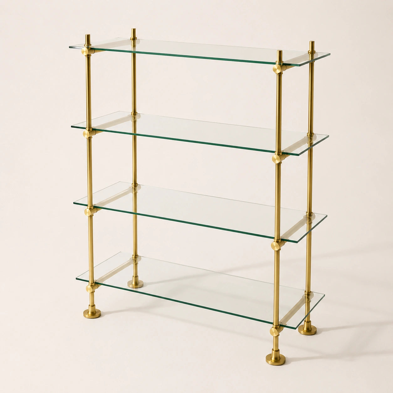 A 4-tier glass shelf bookcase with a polished brass frame, featuring clear glass shelves and round brass finials at the joints and feet, set against a plain off-white background.