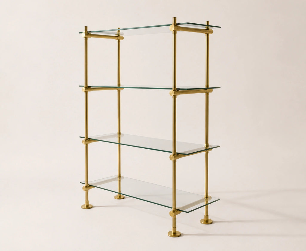 A four-tier glass shelf bookcase with a polished brass frame and round brass feet, photographed against a solid light beige background.