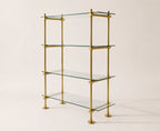 A four-tier glass shelf bookcase with a polished brass frame and round brass feet, photographed against a solid light beige background.