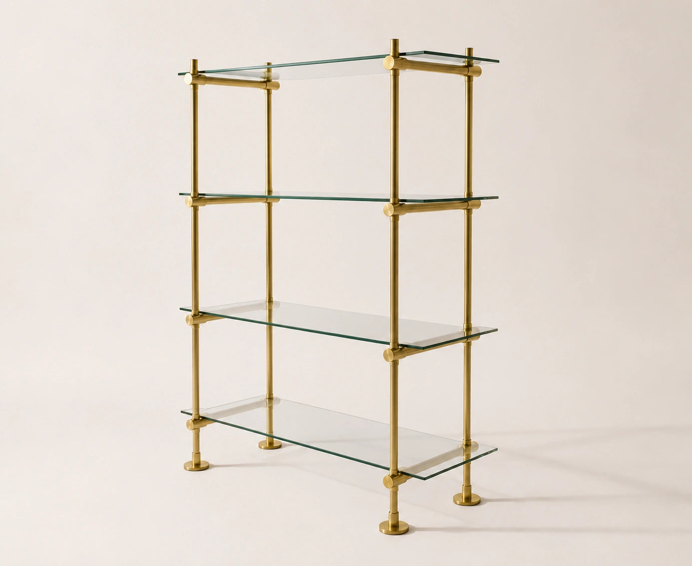 A four-tier glass shelf bookcase with a polished brass frame and round brass feet, photographed against a solid light beige background.