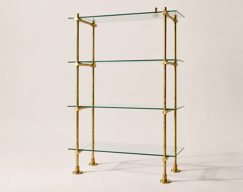 A four-tier glass shelf unit with a polished brass frame, photographed against a solid, warm off-white background.