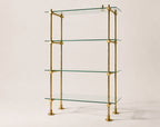 A four-tier glass shelf unit with a polished brass frame, photographed against a solid, warm off-white background.