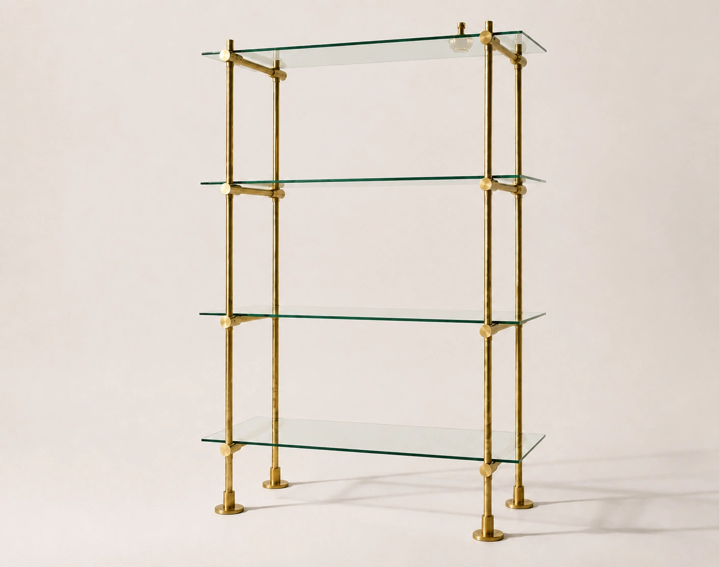 A four-tier glass shelf unit with a polished brass frame, photographed against a solid, warm off-white background.