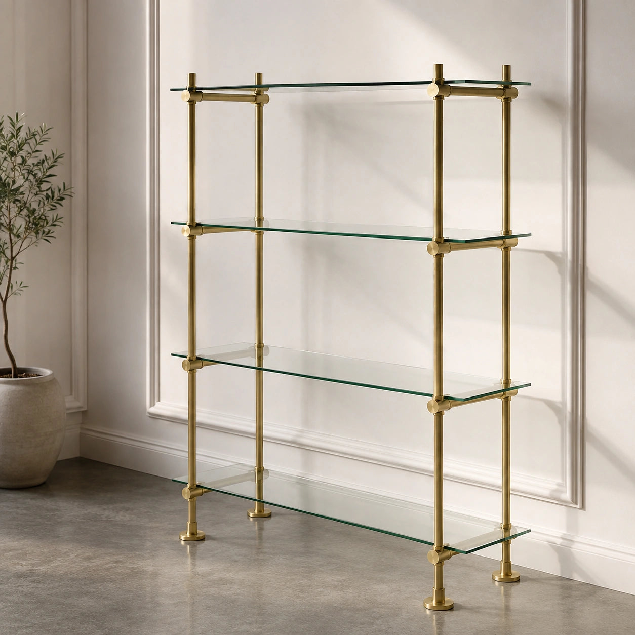 A four-tier brass shelving unit with clear glass shelves, set against a white paneled wall in a styled interior setting.