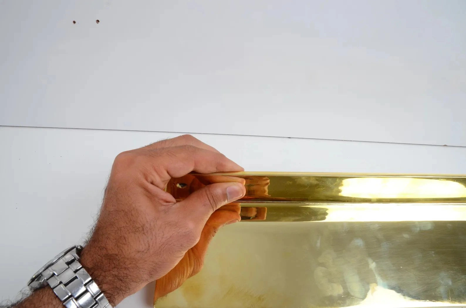 Hand holding a long unlacquered brass floating shelf against a white wall, showcasing polished craftsmanship and reflective golden finish-atlasfinest