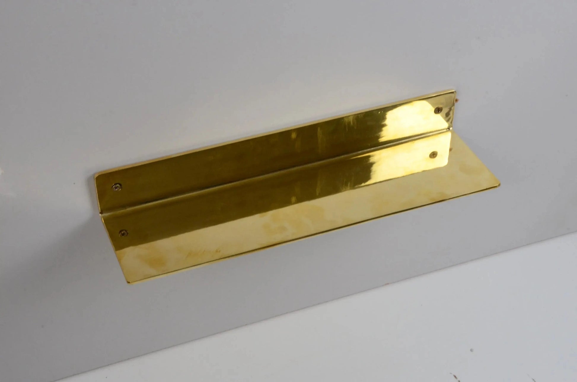 Hand holding a long unlacquered brass floating shelf against a white wall, showcasing polished craftsmanship and reflective golden finish-atlas finest