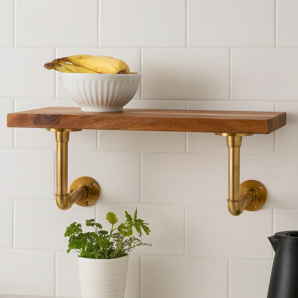 Solid wood floating shelf with two unlacquered brass pipe brackets, white bowl of bananas, small potted herb plant, and modern kitchen background with white subway tiles