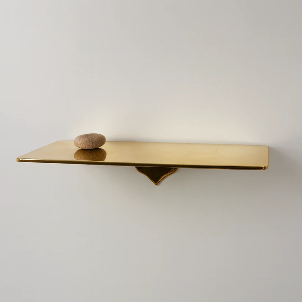 Minimalist unlacquered brass floating shelf with a smooth reflective surface and a small stone on top, mounted on a clean white wall.